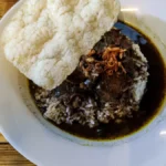 Semangkuk rawon kuah hitam kluwek dengan nasi dan kerupuk udang